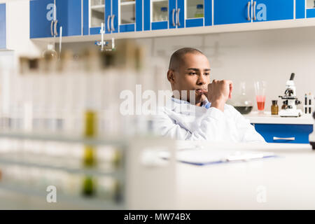 Scienziato maschio durante il lavoro al moderno laboratorio biologico seduta con la mano sul mento Foto Stock