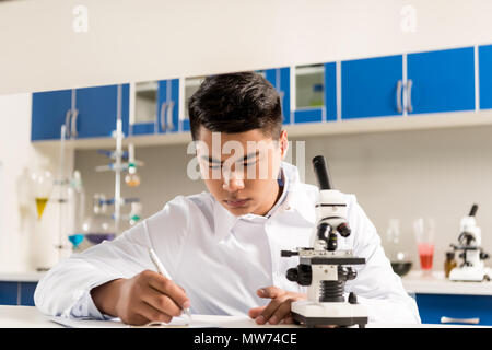 Giovane tecnico di laboratorio in camice bianco prendere appunti su carta dopo aver fatto l'analisi del campione con il microscopio Foto Stock