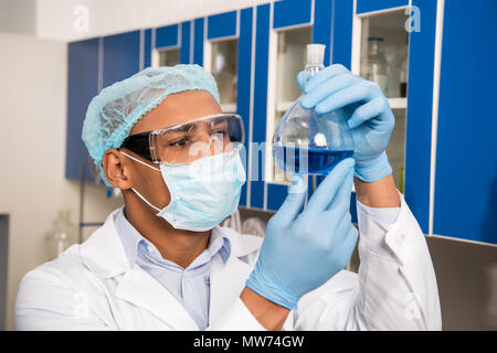 Scienziato in abbigliamento protettivo esaminando tubo di prova con il liquido blu in laboratorio Foto Stock