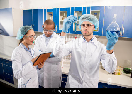 Scienziato in abbigliamento protettivo esaminando le provette con liquido blu in laboratorio con un collega in piedi dietro Foto Stock