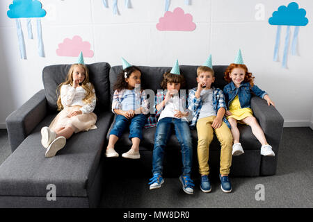 Gruppo di simpatici ragazzi multietnica in party caps seduti sul divano a festa di compleanno Foto Stock