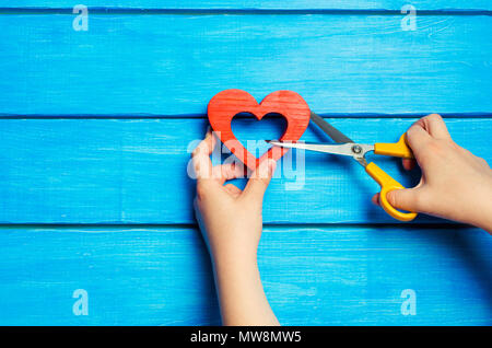 Mani femminili tagliare un di legno cuore rosso con le forbici su uno sfondo blu. Il concetto di rompere le relazioni, litigio, il divorzio. La coppia è divergente. Tradire Foto Stock