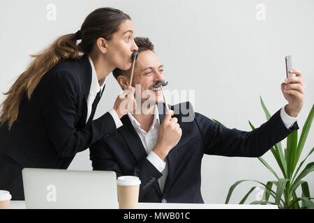 Gentili colleghi smorfie holding baffi finti tenendo selfi Foto Stock