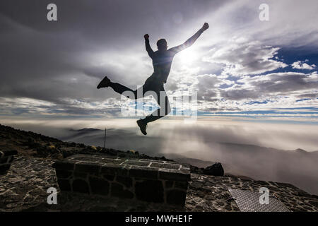 Uomo che salta da banco con le mani in alto di fronte sun con le nuvole in cielo e sulle colline Foto Stock