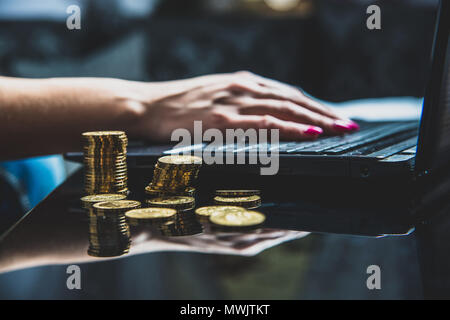 Sul tavolo si trova un sacco di monete d'oro, in background una donna che lavora su un notebook. Home office. La donna lavora sodo per guadagnare denaro, lavorare al compu Foto Stock