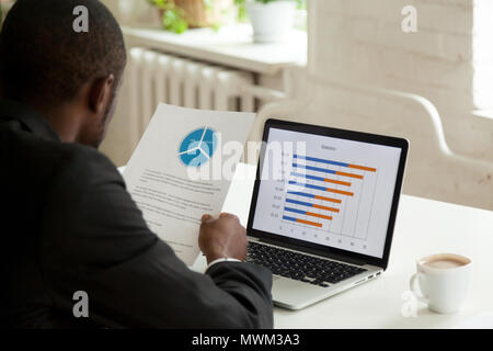 Focalizzato lavoratore nero confrontando i dati finanziari sulla carta e lapto Foto Stock