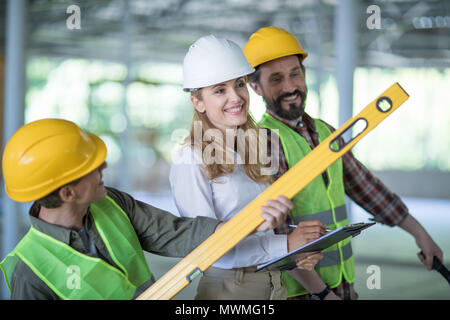 Ispettore sorridente iscritto sulla clipboard mentre in piedi con i lavoratori tenendo lo strumento livello Foto Stock