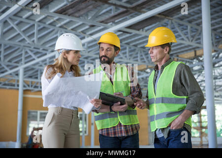 Ispettore seria azienda blueprint e guardando i lavoratori di copricapi rigidi Foto Stock