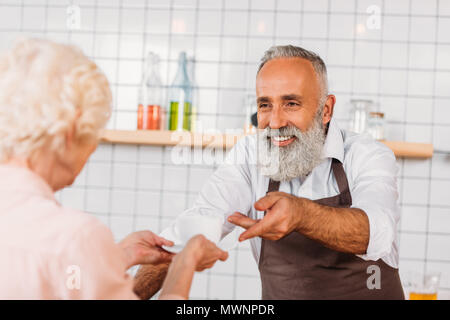 Messa a fuoco selettiva di sorridere senior barista serve tazza di caffè al visitatore Foto Stock