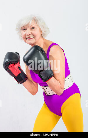 Ritratto di bella sportiva lady senior in guantoni da pugilato sorridente in telecamera isolato su grigio Foto Stock