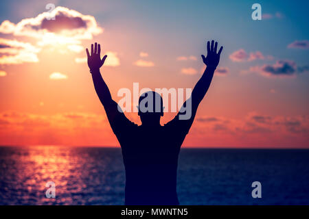 Silhouette di un uomo con alzata fino a mani e gode di fantastiche vista di un tramonto sul mare, godendo le vacanze estive sulla spiaggia Foto Stock