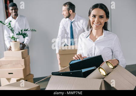 Felice giovane imprenditrice disimballaggio box con forniture di ufficio e sorridente alla telecamera mentre i colleghi di sesso maschile in piedi dietro nel nuovo ufficio Foto Stock