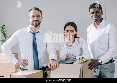 Felice multirazziale colleghi sorridente alla fotocamera e disimballaggio scatole nel nuovo ufficio Foto Stock