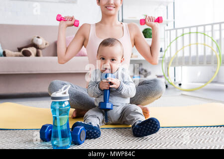 Ritagliato shot di sorridere montare madre facendo esercizi con manubri da seduto sul materassino yoga con il suo bambino a casa Foto Stock