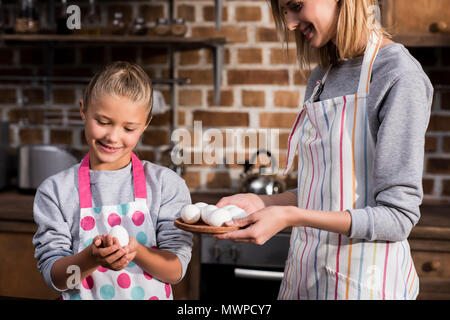 Ritratto di bambina e la madre in grembiuli tenendo il pollo crudo uova durante la cottura insieme Foto Stock