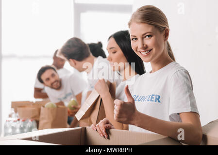 Bellissima femmina volontario che mostra il pollice in su con i colleghi sfocata lavorando sullo sfondo Foto Stock