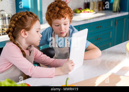 Adorabili bambini nei grembiuli con tavoletta digitale mentre si cucina insieme in cucina Foto Stock