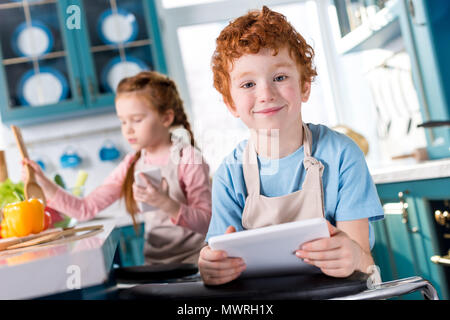 Carino piccolo ragazzo con tavoletta digitale sorridente alla fotocamera mentre il bambino con la cottura dello smartphone dietro Foto Stock