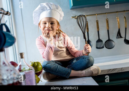 Carino bambino in chef hat holding frusta e sorridente in telecamera in cucina Foto Stock