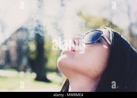 Close-up di un volto di donna soffiando il fumo di una sigaretta. Affascinante ragazza in occhiali da sole. Sottolineato femmina soddisfa il desiderio di nicotina. Concetto di salute, Foto Stock