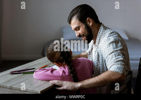 Vista laterale del padre con la figlia piccola seduta a tavola e disegno di album di ritagli a casa Foto Stock
