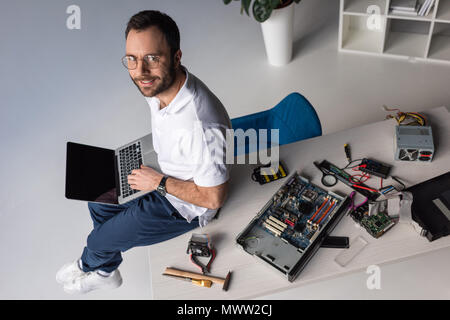 Uomo sorridente seduto sul tavolo con laptop sulle ginocchia Foto Stock