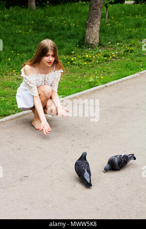 Una bionda a piedi nudi ragazza in un abito bianco alimenta i piccioni. Foto Stock