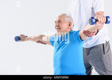 Vista parziale del terapista della riabilitazione assistere senior uomo esercizio con manubri su sfondo grigio Foto Stock