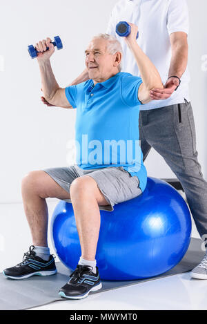 Ritagliato shot del terapista della riabilitazione assistere senior uomo esercizio con manubri sulla palla fitness isolato su grigio Foto Stock