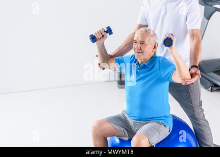 Vista parziale del terapista della riabilitazione assistere senior uomo esercizio con manubri sulla palla fitness su sfondo grigio Foto Stock