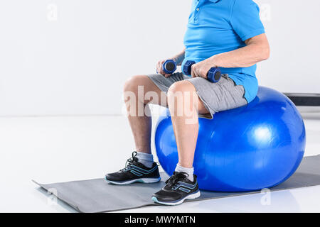 Ritagliato colpo di uomo senior con manubri seduto sulla sfera di fitness su sfondo grigio Foto Stock