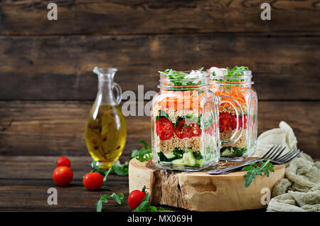 Insalate con la quinoa, rucola, Rafano, pomodori e cetrioli in vasetti di vetro su sfondo di legno. Cibo sano, dieta disintossicante e concetto vegetariano Foto Stock