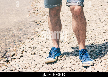 Gambe maschio in tela blu sneakers e blu cut off jeans corti piedi sulla spiaggia di ciottoli vicino all'acqua Foto Stock