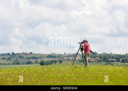 Nei pressi di San Quirico, Toscana, Italia; 10 maggio 2018; donna in piedi in erba Guardando nel mirino della fotocamera reflex digitale su un cavalletto. 2 DSLR sulla sua spalla Foto Stock