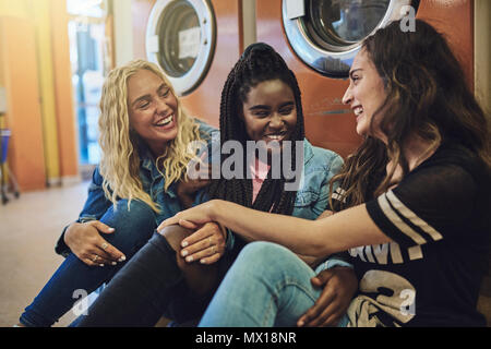 Diversi gruppi di giovani femmine amici seduti su un pavimento di laundromat a parlare e ridere insieme mentre svolgono il servizio lavanderia Foto Stock
