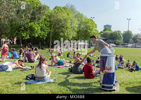 Un giovane uomo mani lattine di Red Bull energy drink rilassanti per gli stranieri in Devonshire verde in Sheffield. Giovani folle seduto su erba, giornata di sole Foto Stock