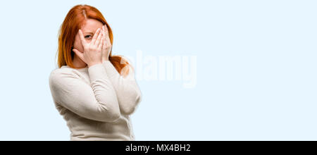 Giovane e bella redhead donna sorridente con sguardo timido spiata attraverso le dita, che copre il viso con le mani cercando confusamente ampiamente isolate su blu Foto Stock