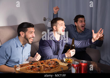 Esaltato amici maschi guardando la tv insieme a casa, godendo di birra e pizza Foto Stock