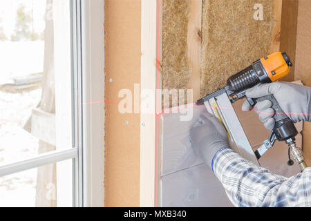Il lavoratore fa opere di finitura delle pareti con un bianco tavola di legno, utilizzando una linea laser livello. Costruzione di isolante termico eco-telaio in legno casa con legno piastre in fibra Foto Stock