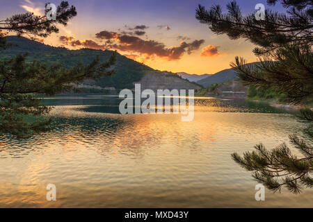 Pino rami framing lago riflettente e ora d'oro tramonto sul lago Zavoj in Serbia, nei pressi di Pirot Foto Stock