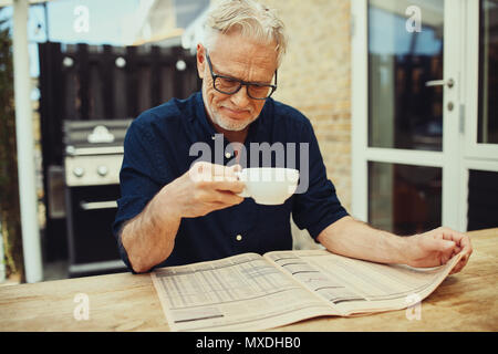 Senior uomo sorridente e tenendo una tazza di caffè mentre seduto fuori al suo patio tabella di lettura di un quotidiano finanziario Foto Stock