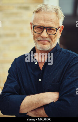 Sorridente senior con barba e con gli occhiali in piedi con le braccia incrociate al di fuori nel suo cortile di casa Foto Stock