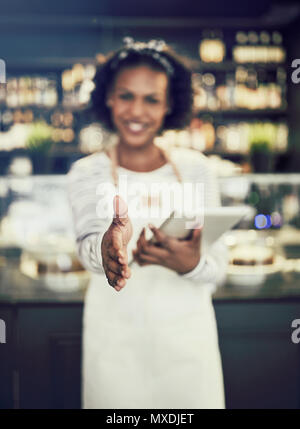 Accogliente giovane africana hostess in piedi in una caffetteria alla moda tenendo un digitale compressa ed estendendo il suo braccio per agitare le mani Foto Stock
