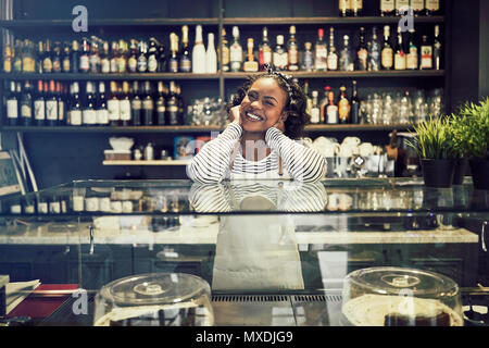 Sorridente giovane imprenditore africano di indossare un grembiule e in piedi dietro il bancone della sua caffetteria alla moda Foto Stock