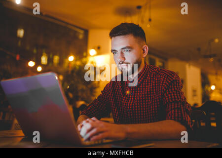 Un bel giovane uomo caucasico con la barba e sorriso toothy in rosso una camicia a scacchi sta lavorando dietro un grigio laptop seduti ad un tavolo di legno. Le mani sulla tastiera. In serata presso la caffetteria Foto Stock