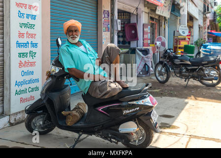 Un uomo anziano in un turbante Sikh, si sta reclinando su una motocicletta di fronte ad un negozio chiuso con un otturatore abbassato . India Giugno 2015 Foto Stock