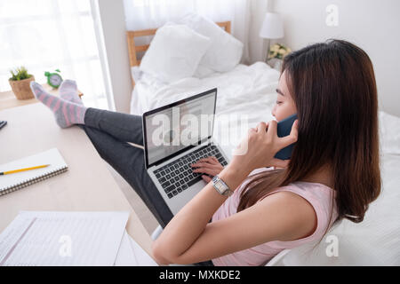 Femmina asiatica libero professionista Parlare su mobile e vedere business grafico su schermo di computer portatile con il resto della gamba sul tavolo in camera da letto a casa.Il lavoro a casa concetto.lavoro fro Foto Stock