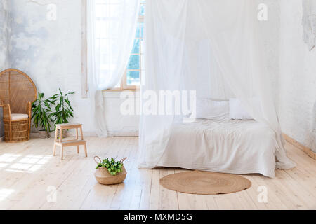 Accogliente camera da letto in colori chiari e con un pavimento in legno, un grande letto a baldacchino, sedia in vimini e cesto di fiori. Semplicità scandinava di design. Eco appartamenti loft. Foto Stock