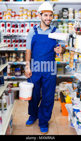 Ridendo operaio in tuta blu con della vernice e spazzole in mani in negozio Foto Stock