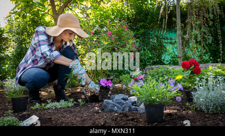 Bellissima femmina giardiniere indossando cappello di paglia piantare fiori nel suo giardino. Concetto di giardinaggio. Giardino paesaggistico proprietario di piccola impresa. Foto Stock